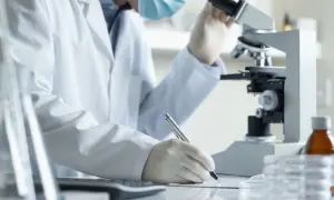 Analityk medyczny w białym fartuchu i masce pracuje przy mikroskopie, notując wyniki. Zastanawiasz się, ile zarabia analityk medyczny?