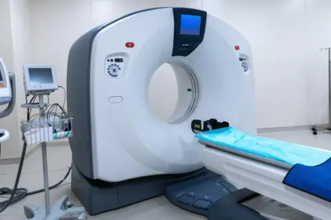Nowoczesny aparat do tomografii komputerowej, gotowy do wykonania badania z kontrastem. Obok stoją monitory i wózek z akcesoriami medycznymi.