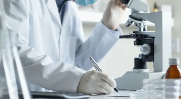 Analityk medyczny w białym fartuchu i masce pracuje przy mikroskopie, notując wyniki. Zastanawiasz się, ile zarabia analityk medyczny?
