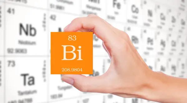 Dłoń trzyma pomarańczową kartkę z symbolem pierwiastka bizmut (Bi) i jego liczbą atomową 83. W tle widać tablicę Mendelejewa.