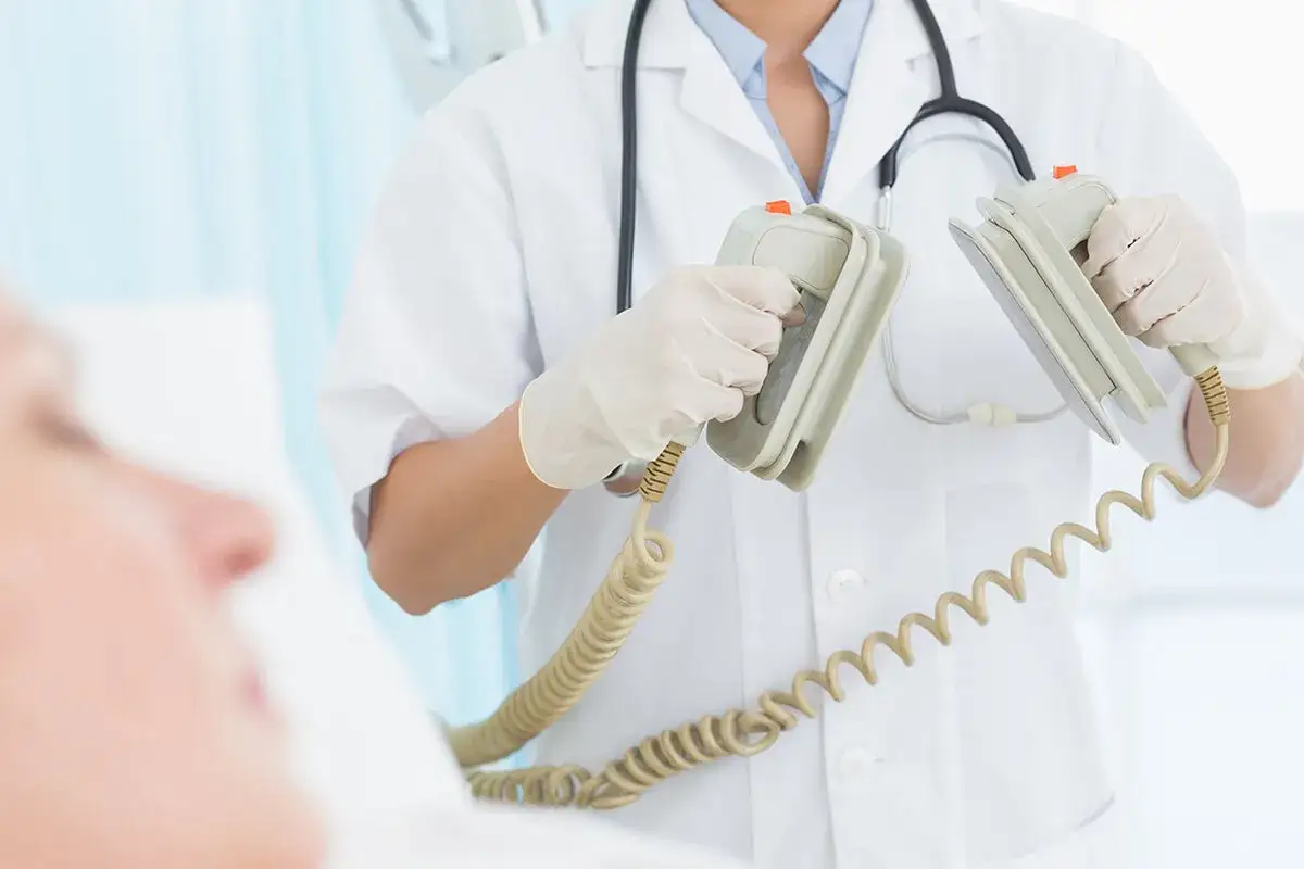 Ratownik medyczny z rękawiczkami trzyma elektrody defibrylatora, gotowy do użycia. Pacjent leży w łóżku. To kluczowy moment, gdzie biosotal może pomóc w przywróceniu funkcji życiowych.
