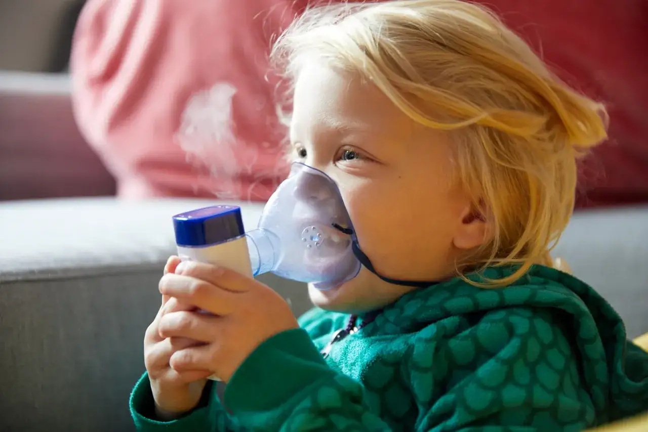 Mała dziewczynka z blond włosami wdycha lek na astmę przez nebulizator. Nie wiadomo na co, ale widać parę.