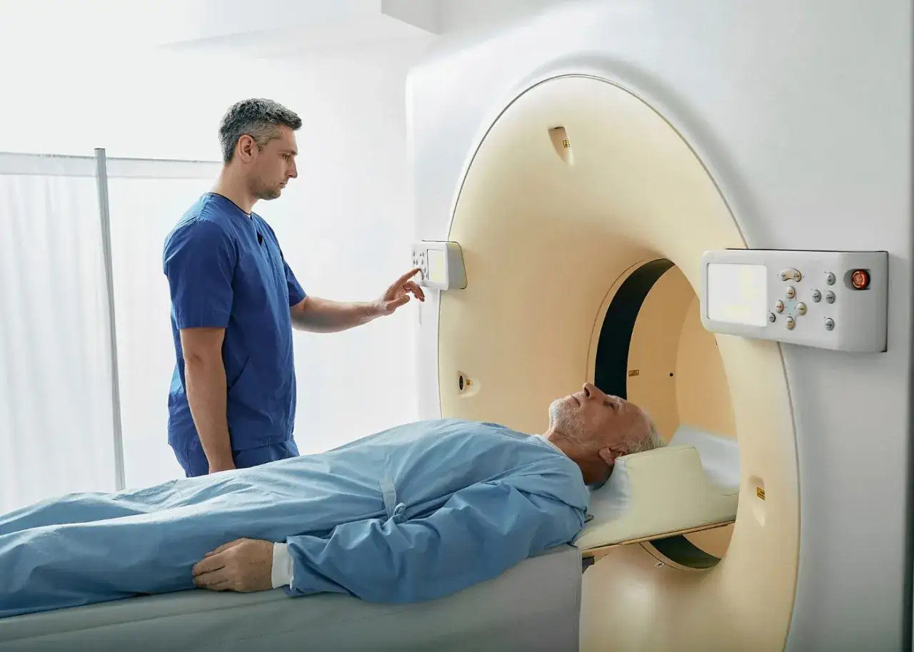 Mężczyzna leży na łóżku podczas badania tomografią z kontrastem. Technik w niebieskim fartuchu obsługuje panel sterowania.